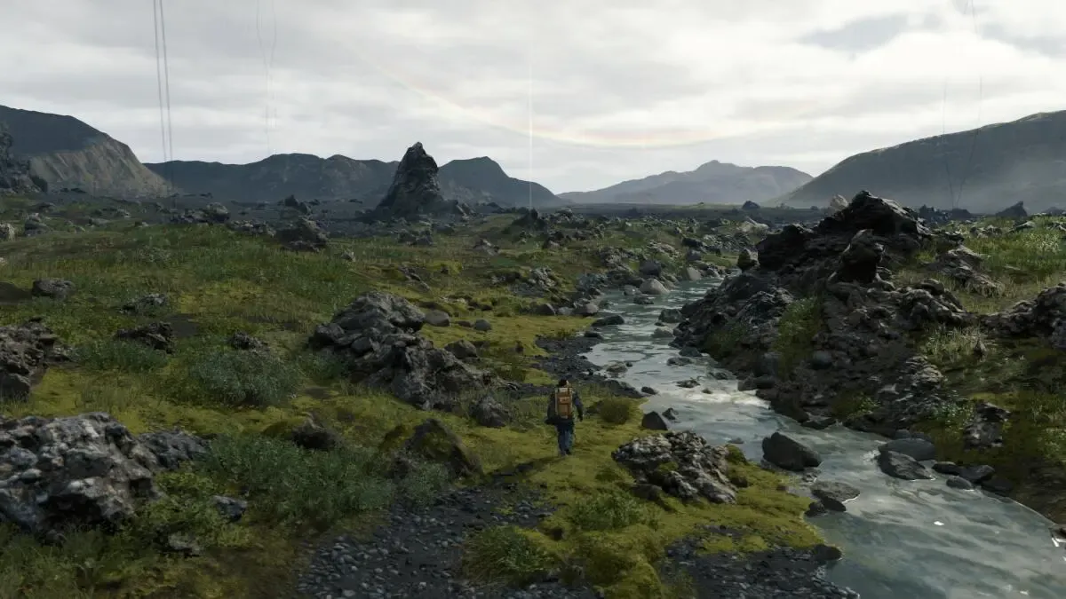 A lone courier with a large backpack walking along a stream through a rocky, mossy valley under a cloudy sky, from Death Stranding